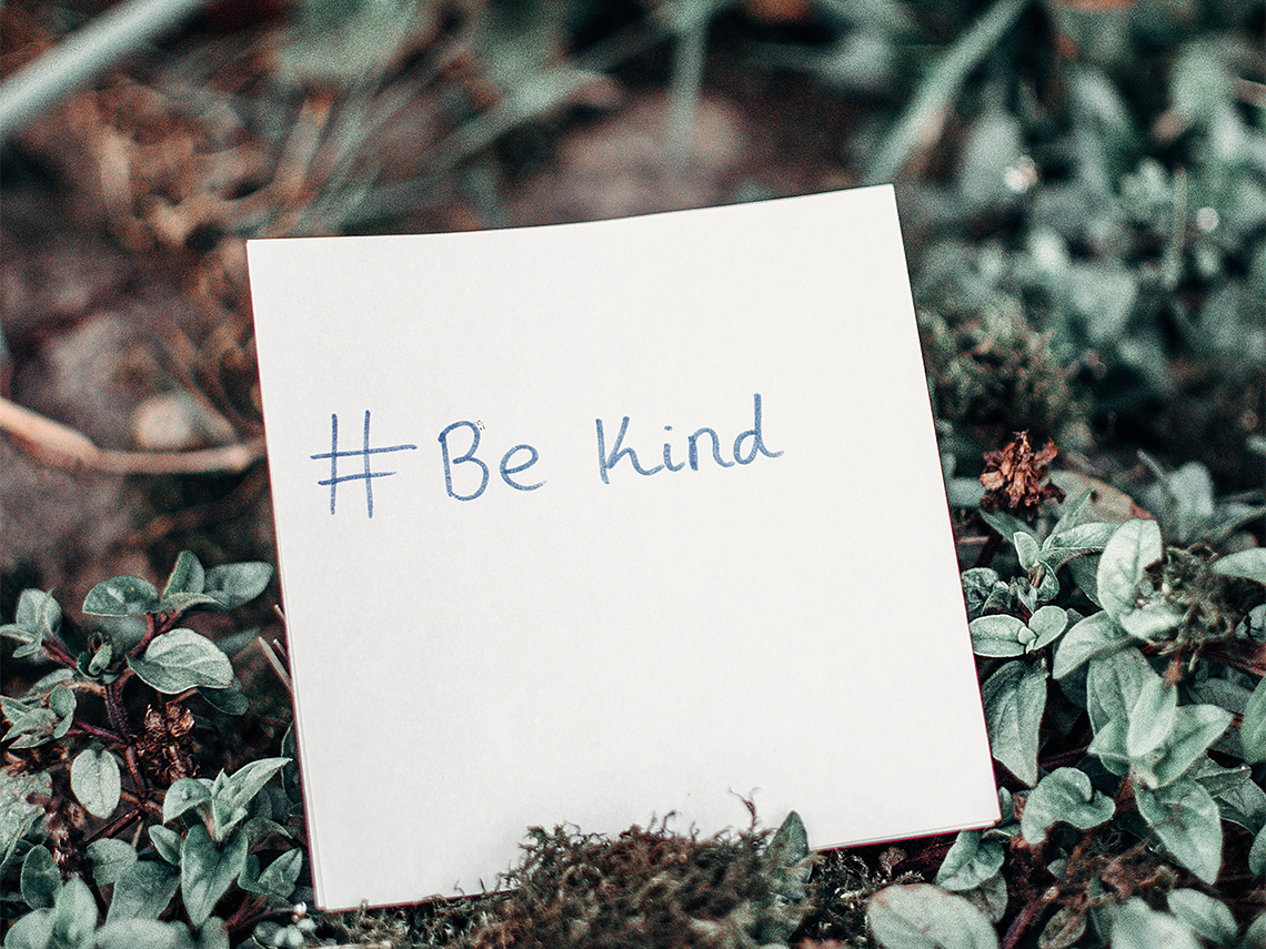 a piece of paper balanced on some leaves on the ground with '#be kind' written in blue lettering